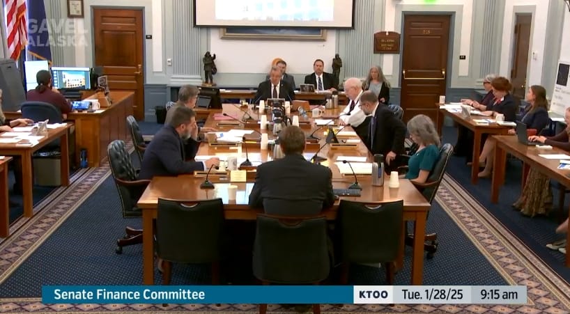 A wide shot of the Senate Finance Committee with people in suits sitting around a long wide meeting table flanked by smaller tables of each legislators' aides.